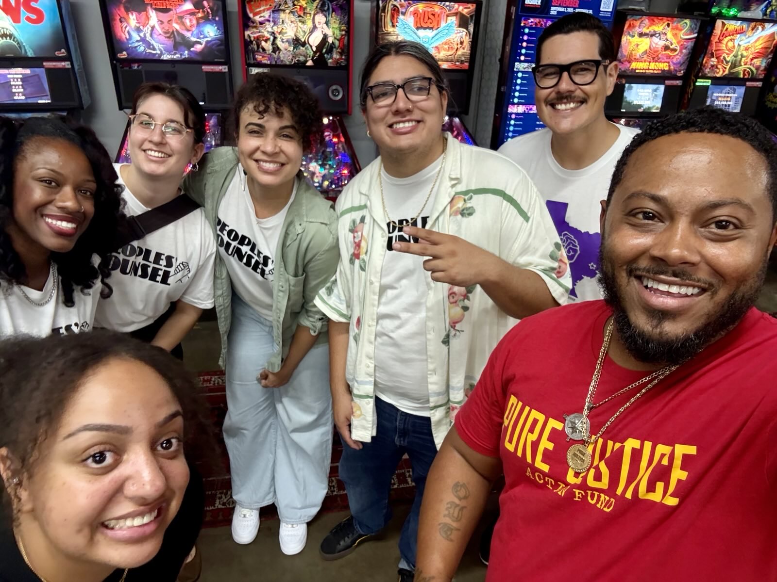 A group of seven diverse individuals posing together in front of arcade machines, all smiling and wearing themed t-shirts. The scene is lively and colorful, showcasing a fun atmosphere.